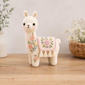 Könnte beinhalten: Eine cremefarbene gehäkelte Lama-Figur mit gestickten pastellfarbenen Blumendetails am Hals und Bein. Das Lama trägt eine dekorative Satteldecke mit Blumenmuster und kleinen cremefarbenen Quasten an der unteren Kante.