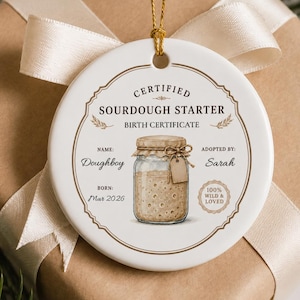 May include: A round white ceramic ornament featuring a printed sourdough starter birth certificate design. The text reads Certified Sourdough Starter Birth Certificate, Name Doughboy, Adopted By Sarah, Born Mar 2026, and 100% Wild & Loved.
