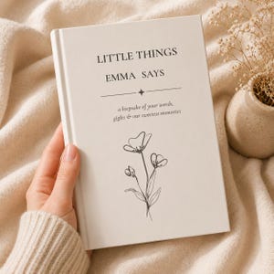 Peut inclure: Un livre à couverture rigide de couleur crème avec un texte noir indiquant LITTLE THINGS EMMA SAYS, a keepsake of your words, giggles & our sweetest memories, orné d'un dessin au trait noir représentant une fleur à trois tiges.