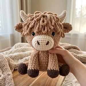 May include: A crocheted Highland cow plushie in shades of tan and brown. The plush toy has a light tan muzzle, horns, and inner ears, with dark brown hooves. The cow has a fluffy, curly mane and a friendly expression.