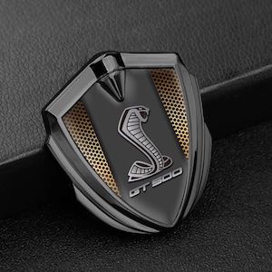 May include: A black and silver emblem with a shield shape. The emblem features a cobra design in silver, set against a black background with gold mesh accents. The text "GT 500" is at the bottom.