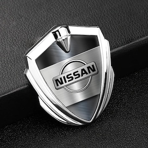 Peut inclure: Un emblème Nissan chromé en forme de bouclier. Il présente un centre en métal brossé avec le logo Nissan dans un rectangle noir. Le dessus est pointu et l'ensemble est très brillant.