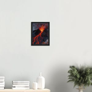 Könnte beinhalten: Ein gerahmter Wandkunstdruck, der einen Vulkanausbruch zeigt, bei dem leuchtend orangefarbene und rote Lava die dunklen, zerklüfteten Hänge eines Berges hinunterfließt. Die Szene zeigt dicke graue Rauch- und Aschewolken über dem Gipfel.