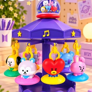 Op de afbeelding: Een paars carrousel-speeltje met BT21-figuren. De bovenkant heeft een doorzichtige koepel met miniatuurfiguurtjes. Vijf hangende karakterbedels in geel, lichtpaars, rood, blauw en roze draaien rond een centrale paarse zuil op een geschulpte voet.
