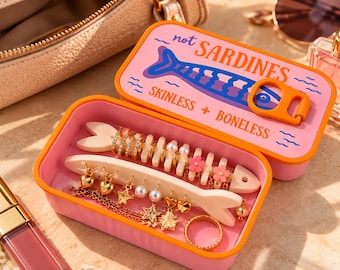 Op de afbeelding: Een roze rechthoekig sieradendoosje met een oranje rand, voorzien van een blauwe visafbeelding en de tekst "not SARDINES SKINLESS + BONELESS" op het deksel. Binnenin bevinden zich twee witte visvormige oorbelhouders met goudkleurige oorbellen in de vorm van hartjes, sterren, parels en roze bloemen, plus een gouden ring.