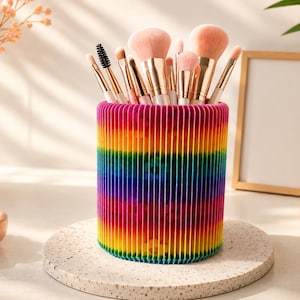 May include: A cylindrical make-up brush holder featuring a vibrant rainbow-coloured accordion-style pleated exterior. The holder is filled with various make-up brushes with light pink bristles and metallic gold ferrules, standing upright in a cluster.