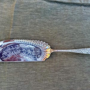May include: A silver-colored serving utensil with a wide, curved blade and a long, decorated handle. The blade features an etched floral design. The edge of the blade has a decorative, scalloped design. The handle is also decorated with an etched pattern.