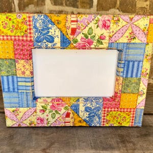 May include: A rectangular photo frame with a patchwork design. The frame is made of fabric squares in various colors, including yellow, pink, blue, and red, with floral and geometric patterns. The frame has a white rectangular opening.