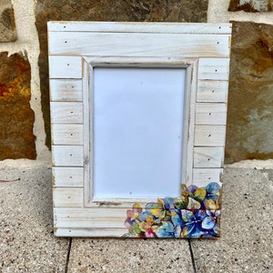 Könnte beinhalten: Weißer Holzbilderrahmen mit einer abgenutzten Oberfläche und einem Blumendesign in der rechten unteren Ecke. Der Rahmen hat einen weißen Passepartout und eine klare Glasscheibe.