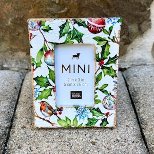 May include: A rectangular mini picture frame with a Christmas theme. The frame is decorated with holly, ornaments, and a bird. The frame is 2 inches by 3 inches (5 cm x 7.6 cm) and has the word "MINI" printed on the front.