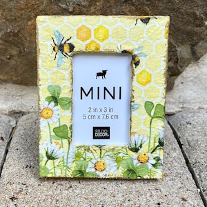 May include: A small yellow picture frame with a honeycomb pattern and bee illustrations. The frame is decorated with white daisies and green leaves. The frame measures 2 inches by 3 inches or 5 cm by 7.6 cm. The text "MINI" is printed on the frame.