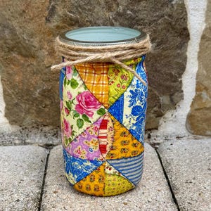 May include: A glass jar with a patchwork design featuring floral, geometric, and striped patterns in shades of blue, yellow, red, and green. The jar is tied with twine around the lid.