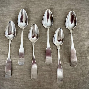 May include: Six antique silver spoons arranged on a textured, neutral-toned surface. The spoons have a reflective, polished finish, with elongated bowls and slender handles. The spoons are likely for serving or dining.