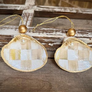 May include: Two decorative ornaments crafted from oyster shells, each featuring a hand-painted white and tan checkered pattern on the interior. The edges are trimmed in gold, and each shell is topped with a small white pearl and a gold bead.