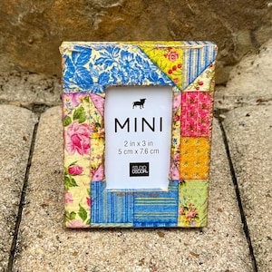May include: A small, colorful patchwork picture frame with a white mat and the words "MINI 2 in x 3 in 5 cm x 7.6 cm STUDIO DECOR." printed on it.