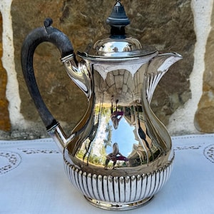 May include: A silver-plated coffee pot with a dark wooden handle and lid finial. The pot has a ribbed design around the base and a spout for pouring. The reflective surface mirrors the surroundings.