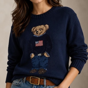 May include: A navy blue long-sleeve crewneck sweater featuring a knitted graphic of a teddy bear wearing a dark sweater with an American flag design, blue jeans, and brown shoes. The sweater has ribbed cuffs and a ribbed hem at the neckline.
