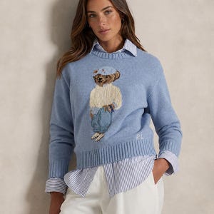 May include: A light blue long-sleeve knit sweater featuring a central intarsia-knit bear graphic wearing a cap, cream sweater, and blue pants. The sweater has a ribbed crew neckline, cuffs, and hem, with the letters RL embroidered in white.