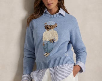 May include: A light blue long-sleeve knit sweater featuring a central intarsia-knit bear graphic wearing a cap, cream sweater, and blue pants. The sweater has a ribbed crew neckline, cuffs, and hem, with the letters RL embroidered in white.