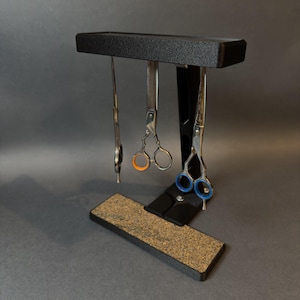 Peut inclure: Un support noir avec une base rectangulaire et une barre supérieure horizontale conçue pour maintenir des ciseaux. Trois paires de ciseaux de toilettage en métal pendent de la barre, avec des anneaux orange, bleus et argentés.