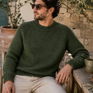 Puede incluir: Un suéter de manga larga en color verde oliva oscuro con una textura de punto grueso, cuello redondo acanalado y puños acanalados. La prenda está confeccionada con mangas raglán y un patrón de tejido denso y resistente de color uniforme.
