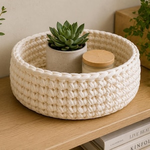 Op de afbeelding: Een ronde, gebroken witte gehaakte mand met een dik, gestructureerd steekpatroon. In de mand staat een kleine groene vetplant in een grijze pot en een kaars met een licht houtkleurig deksel, gemaakt van dik, zacht koordmateriaal.