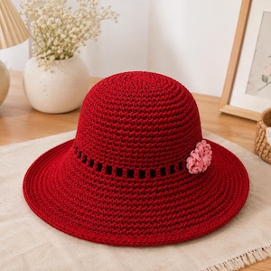 May include: A deep red crochet sun hat featuring a wide brim and a textured crown. The hat includes a decorative row of open square cutouts around the base of the crown and a small, light pink crocheted flower accent attached to the side.