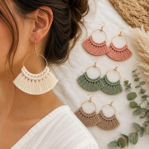 May include: A collection of macrame hoop earrings featuring gold-toned metal rings adorned with knotted cotton cord in a fan-shaped fringe design. The set includes pairs in cream, dusty rose, sage green, and tan, each with a simple hook closure.