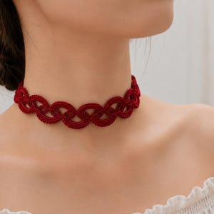 May include: A deep red choker necklace featuring an intricate, repeating woven or knotted pattern. The piece is constructed from a textured thread material, forming a continuous series of interconnected loops that sit snugly around the neck.