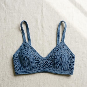 Op de afbeelding: Een stoffig blauwe gehaakte bralette met een driehoekig cupontwerp met ingewikkelde opengewerkte steken, een stevige basisband en twee dikke schouderbandjes. Het kledingstuk is gemaakt van garen met een zichtbare gebreide textuur.