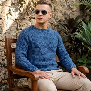 Könnte beinhalten: Ein blauer Langarmpullover mit strukturiertem Strickmuster, Rundhalsausschnitt und gerippten Bündchen. Der Pullover hat eine Raglan-Ärmelkonstruktion und wird mit einer hellbeigen Hose und einer dunkel getönten Pilotenbrille getragen.