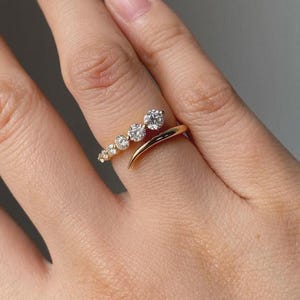 May include: A gold ring featuring a series of graduated, round-cut diamonds. The ring's design curves around the finger, with the diamonds set in a line. The ring is shown on a hand, highlighting the jewelry's sparkle and design.