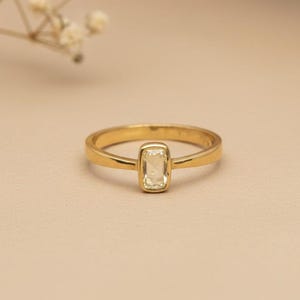 May include: A gold ring featuring a single rectangular, light yellow gemstone set in a smooth bezel setting. The band is polished gold with a simple, rounded profile, showcasing a clean design that highlights the central stone's clarity and color.