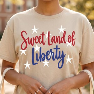 Puede incluir: Camiseta beige con la frase "Sweet Land of Liberty" en letras rojas y azules, rodeada de estrellas blancas. La camiseta de manga corta la lleva una persona que sostiene una bolsa de mano.
