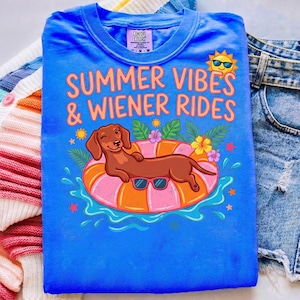 以下が含まれることがあります： 「SUMMER VIBES & WIENER RIDES」の文字と、サングラスをかけたダックスフンドの漫画が描かれた青いTシャツ。デザインには太陽、花、水しぶきが含まれています。