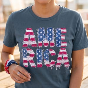 Op de afbeelding: Een leisteenblauw t-shirt met het woord "AMERICA" in een distressed, patriottisch ontwerp. De letters zijn gemaakt om op de Amerikaanse vlag te lijken, met rode en witte strepen en blauwe velden met witte sterren. Een bijpassende armband wordt om de pols gedragen.
