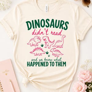 May include: A cream-colored short-sleeve t-shirt featuring the dark green text DINOSAURS and HAPPENED TO THEM, with pink text didn't read and we know what. The design includes five pink dinosaur illustrations and small green grass accents.