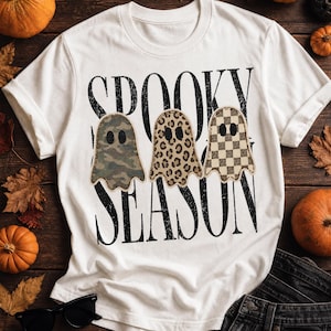 May include: A white short-sleeve t-shirt featuring the black text SPOOKY SEASON and three ghost illustrations. The ghosts have distinct patterns: one with green camouflage, one with a brown leopard print, and one with a tan checkerboard pattern.