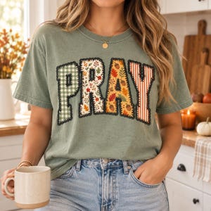 Könnte beinhalten: Ein salbeigrünes Kurzarm-T-Shirt mit dem Wort PRAY in großen Blockbuchstaben. Jeder Buchstabe hat ein eigenes Muster: grünes Vichy-Karo, Herbstblätter, Eicheln mit Herbstlaub sowie orange-weiße vertikale Streifen mit einer Ziernaht.