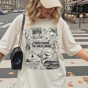 May include: An off-white short-sleeve t-shirt featuring a black graphic print with the text Noah Kahan The Great Divide, surrounded by illustrations of a telephone, a window, a lantern, a ghost, a bird, and various song lyrics in a collage style.