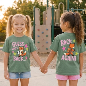 Puede incluir: Una camiseta verde salvia con un zorro de dibujos animados con mochila y cuaderno. El frente muestra el texto rosa "GUESS WHO'S BACK..." con estrellas amarillas y rosas. La parte posterior muestra al zorro con un lápiz y el texto rosa "BACK AGAIN!"