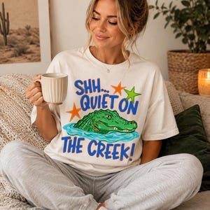 May include: A cream-colored short-sleeve t-shirt featuring a graphic of a green alligator in blue water with orange and green starfish. The text reads SHH... QUIET ON THE CREEK! in blue letters. The shirt is paired with light gray sweatpants.