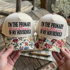 May include: Two off-white baseball caps featuring the black embroidered text I'M THE PRIMARY and OF MY HOUSEHOLD with the pink cursive word breadloser in the center. The brims are decorated with a colorful floral pattern of red and yellow roses.