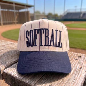 May include: A baseball cap with a cream-colored crown featuring thin vertical navy blue pinstripes and the word SOFTBALL embroidered in navy blue block letters across the front. The cap has a solid navy blue brim with visible stitching.