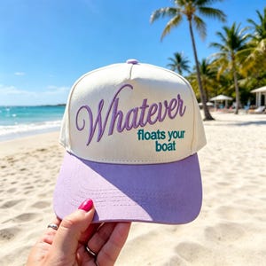 May include: A cream-colored baseball cap with a purple brim and purple embroidered text that reads "Whatever" in a cursive script, followed by the teal printed text "floats your boat" underneath. The cap features a structured front panel.