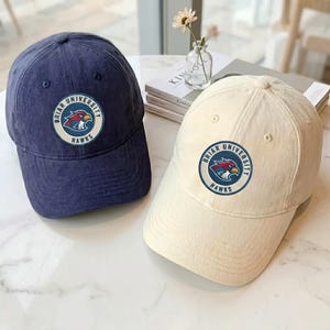 May include: Two baseball caps, one navy blue and one cream, each featuring a circular patch with the text Briar University Hawks and a hawk mascot graphic. The caps have a structured crown, a curved brim, and stitched eyelets on the panels.