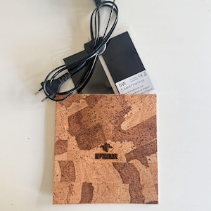 Peut inclure: Un coussin chauffant carré en liège avec une texture brune marbrée et un petit logo noir au centre. Il est muni d'un cordon d'alimentation noir et d'un élément chauffant en plastique transparent avec une étiquette blanche indiquant 5W, 14x15cm.