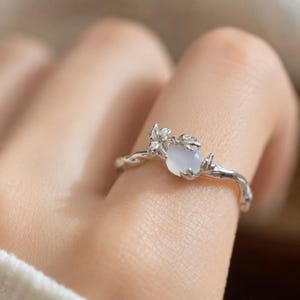 May include: A silver-toned ring featuring a band designed to resemble a twisted vine. The ring is adorned with a small, smooth, translucent pale blue cabochon stone and a delicate metal flower detail positioned next to the stone.
