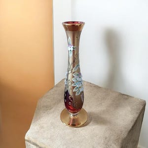 Peut inclure: Un vase soliflore en verre haut et élancé avec une finition bronze métallique et une base rouge profond. Le vase présente des motifs floraux peints à la main en blanc, bleu et vert, rehaussés par de délicats motifs de volutes dorées.