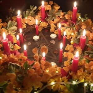 May include: A circular wreath decorated with orange and yellow flowers, green leaves, and small white string lights. Multiple red taper candles and small white tea light candles are arranged around a central dark surface with a pentagram symbol.